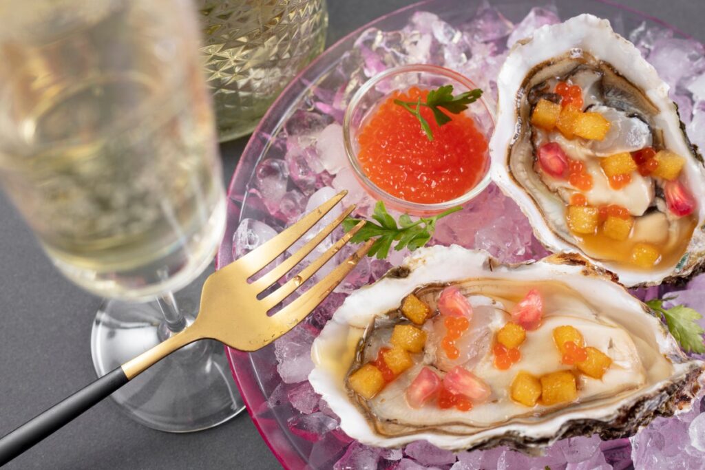 Oysters on crushed ice with orange roe, pomegranate seeds and yellow garnish beside a glass and gold fork
