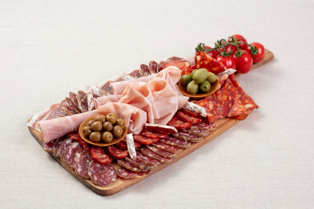 A wooden board holds Spanish-style cured meats, folded ham, olives, cherry tomatoes, and sliced sausage.
