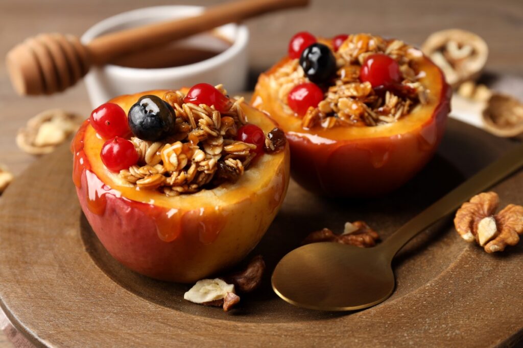 Two baked apples filled with granola, berries and honey sit beside a spoon, walnuts and a honey dipper.