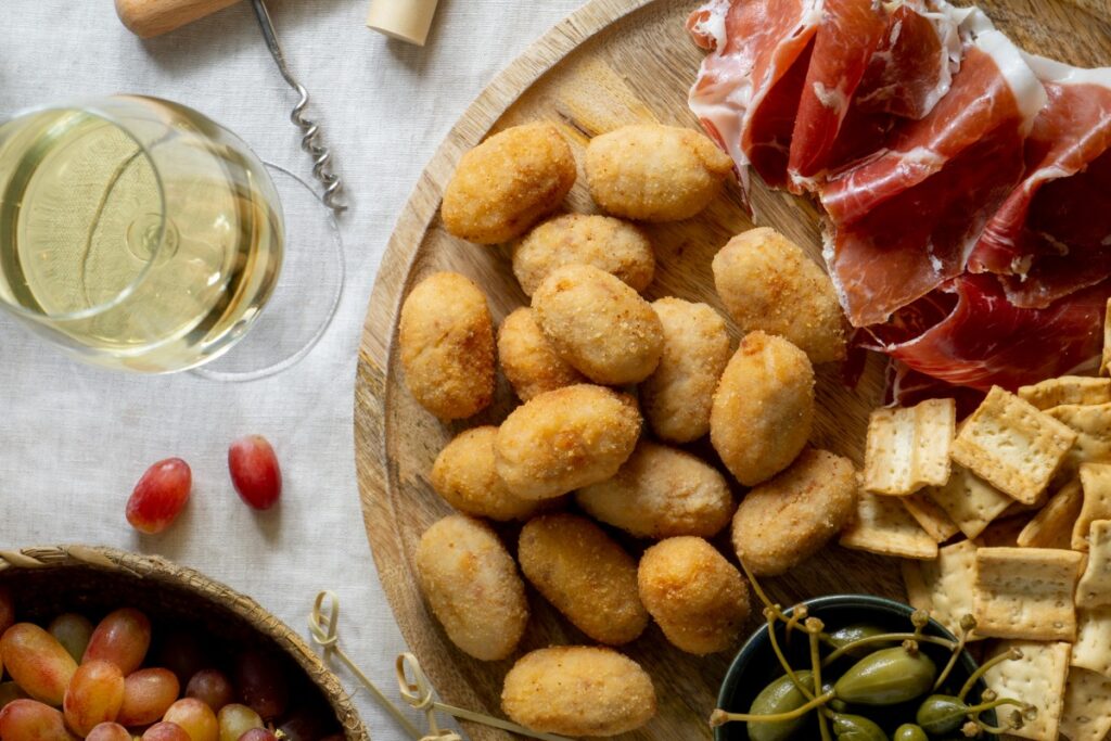 Spanische Kroketten, Serranoschinken, Cracker und Oliven auf einer rustikalen Servierplatte neben einem Glas Weißwein