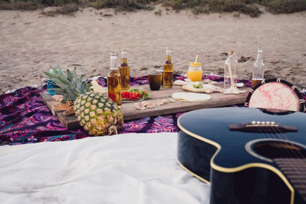 A guitar rests beside a beach picnic with pineapple, drinks, flowers, and a rustic wooden table on a patterned blanket.