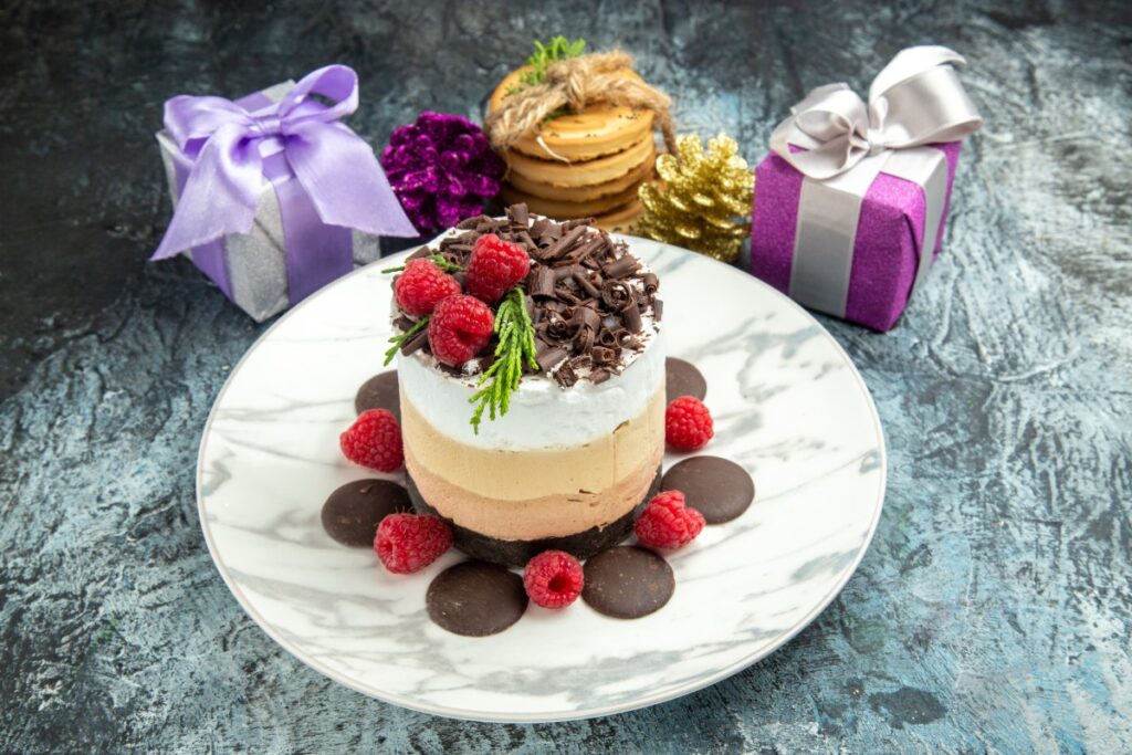 A round cheesecake topped with raspberries and chocolate curls, surrounded by chocolate discs and Christmas gifts.