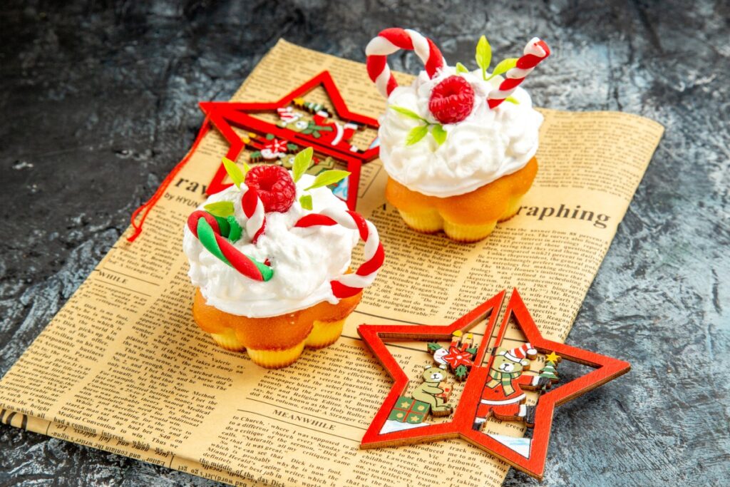 Two cupcakes topped with whipped cream, red berries and candy canes rest on vintage newspaper pages with red star ornaments.