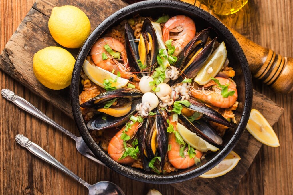A Spanish seafood paella fills a black pan with mussels, prawns, squid, lemon wedges, and herbs on a rustic board