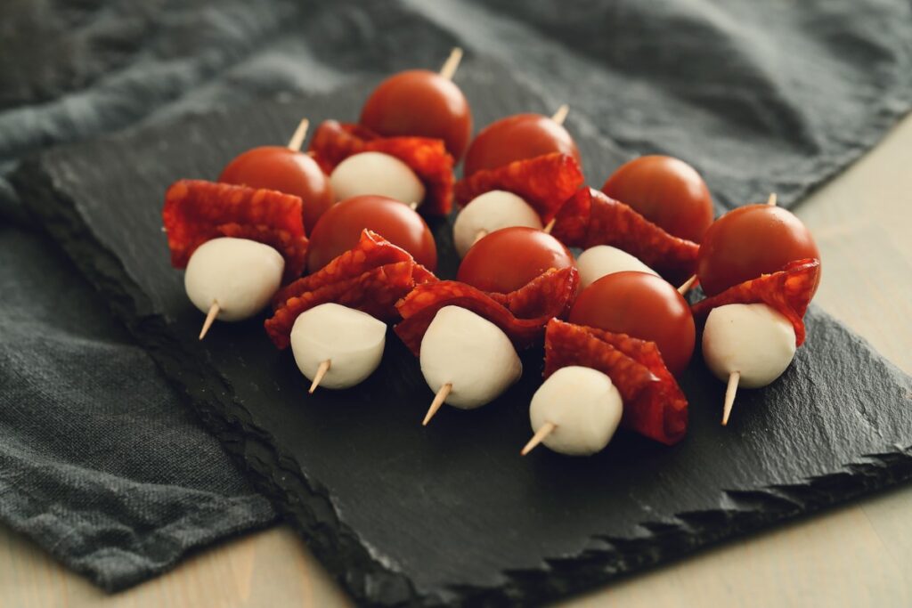 Skewers of red cured meat, cherry tomatoes, and white mozzarella balls arranged on a dark slate serving board.