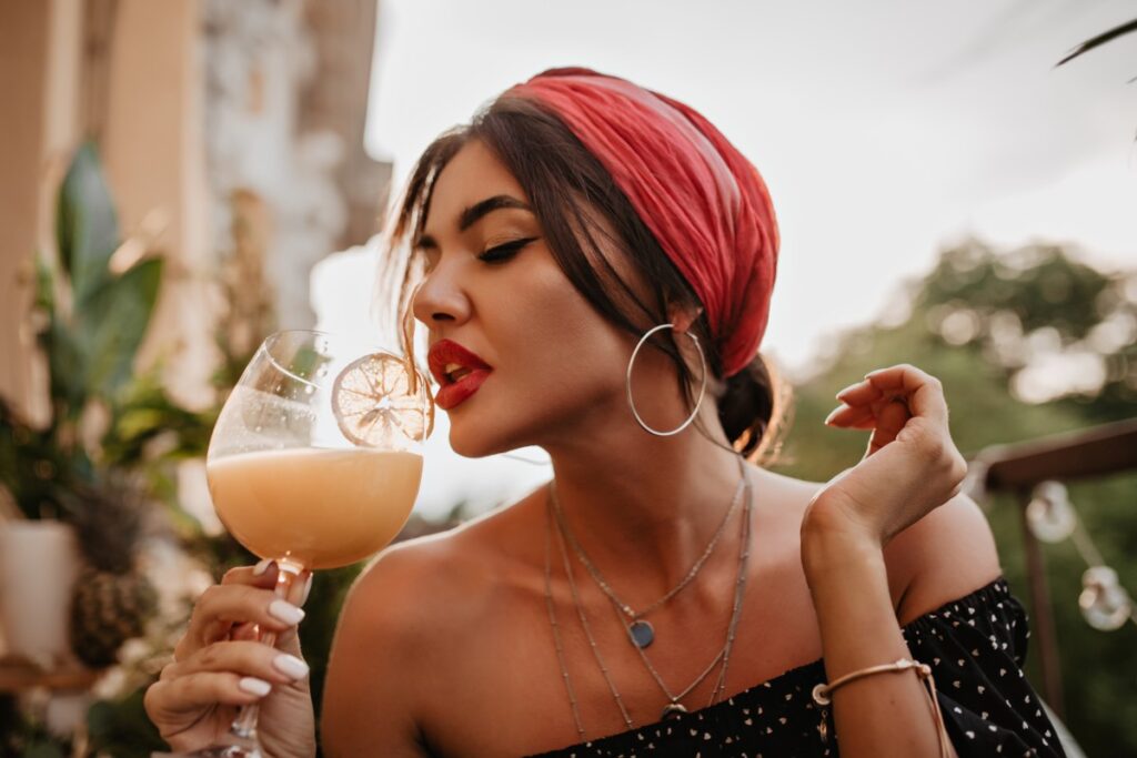 A woman in a red headwrap and black off-shoulder top holds a creamy cocktail outdoors beside leafy plants.