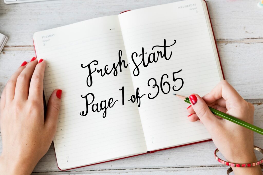 Offenes Notizbuch mit dem Schriftzug „Fresh start, Page 1 of 365“ und Händen mit rotem Nagellack auf einem Holztisch
